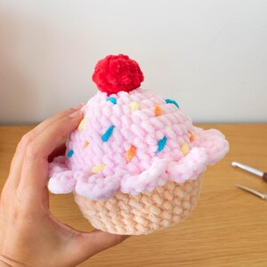 Op de afbeelding: Een handgemaakte gehaakte cupcake met roze glazuur, kleurrijke sprinkles en een rode pompon. De cupcake heeft een lichtbruine basis en wordt door een hand vastgehouden. Het Nomad Knot-logo is zichtbaar.