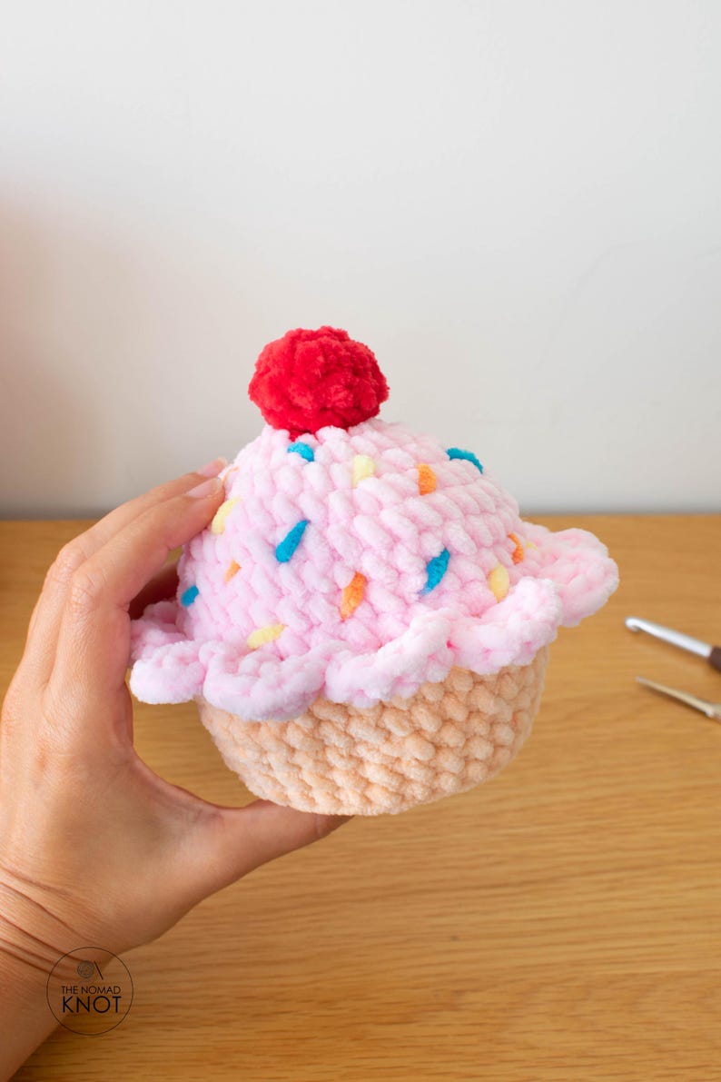 Op de afbeelding: Een handgemaakte gehaakte cupcake met roze glazuur, kleurrijke sprinkles en een rode pompon. De cupcake heeft een lichtbruine basis en wordt door een hand vastgehouden. Het Nomad Knot-logo is zichtbaar.