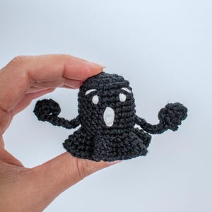 May include: A black crocheted ghost with white eyes and a mouth, holding out its arms. The ghost is being held in a hand.