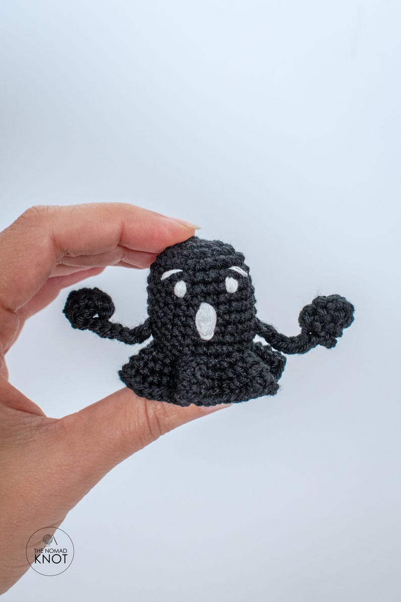 May include: A black crocheted ghost with white eyes and a mouth, holding out its arms. The ghost is being held in a hand.