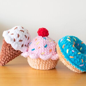 Op de afbeelding: Drie gehaakte dessertartikelen: een ijshoorntje met witte en bruine hagelslag, een roze cupcake met hagelslag en een rode kers, en een blauwe donut met kleurrijke hagelslag. De items zijn gemaakt van zacht garen en liggen op een houten oppervlak.