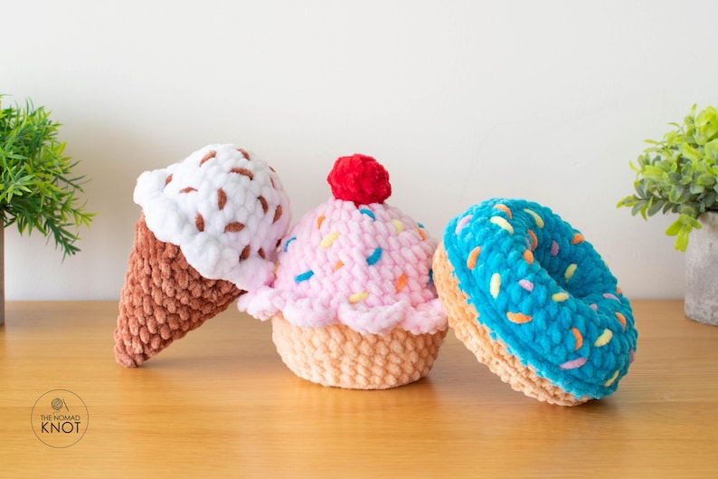 Op de afbeelding: Drie gehaakte dessertartikelen: een ijshoorntje met witte en bruine hagelslag, een roze cupcake met hagelslag en een rode kers, en een blauwe donut met kleurrijke hagelslag. De items zijn gemaakt van zacht garen en liggen op een houten oppervlak.