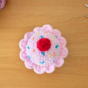Op de afbeelding: Een handgemaakte gehaakte cupcake met een roze basis en glazuur. De glazuur is versierd met kleurrijke hagelslag en een rode kers. Op een houten oppervlak, met garen en knutselgereedschap in de buurt.