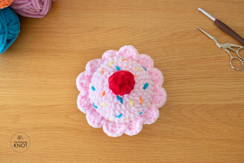 Op de afbeelding: Een handgemaakte gehaakte cupcake met een roze basis en glazuur. De glazuur is versierd met kleurrijke hagelslag en een rode kers. Op een houten oppervlak, met garen en knutselgereedschap in de buurt.