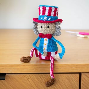 May include: A crocheted Uncle Sam doll in red, white, and blue. The doll has a top hat, a bow tie, and a blue jacket with white stars. The legs are striped red and white, and the feet are brown. The doll is sitting on a wooden surface.