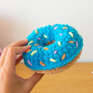 May include: A handmade, plush crochet donut in a vibrant blue color, adorned with colorful sprinkles. The donut has a light brown bottom and a hole in the center. Other yarn balls are visible in the background.