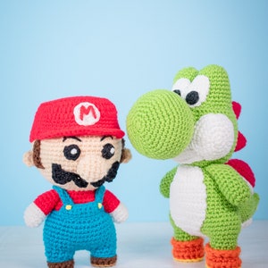 May include: Crocheted plush toys of Mario and Yoshi. Mario is wearing a red hat with an 'M' on it, a red shirt, and blue overalls. Yoshi is green with a white belly and orange shoes.