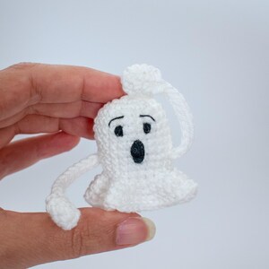 May include: White crocheted ghost with black eyes and a mouth, holding its head with one hand. The ghost is small and is being held by a hand.