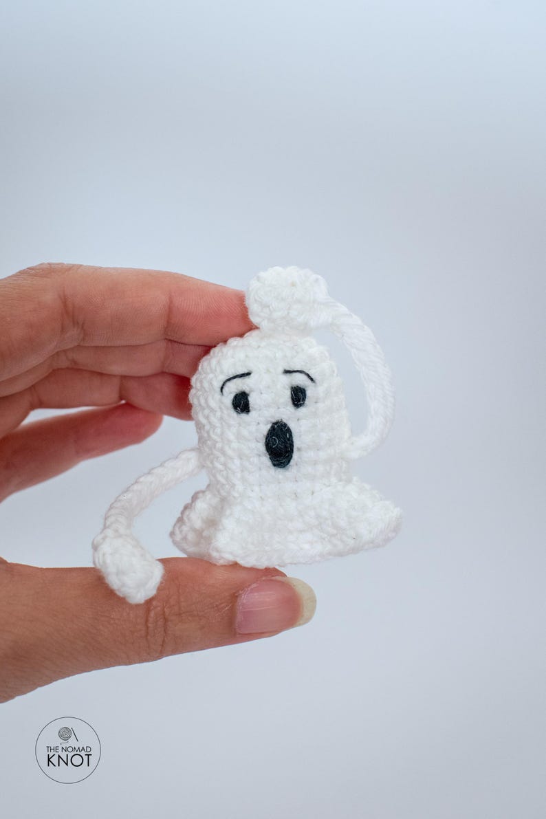 May include: White crocheted ghost with black eyes and a mouth, holding its head with one hand. The ghost is small and is being held by a hand.