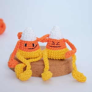 May include: Two crocheted candy corn characters with different facial expressions. The candy corn on the left is smiling, while the candy corn on the right is frowning. Both are sitting on a cork surface.
