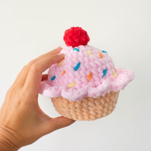 Op de afbeelding: Een handgemaakte gehaakte cupcake met roze glazuur, kleurrijke sprinkles en een rode kers. De cupcake heeft een lichtbruine, getextureerde basis en wordt in een hand gehouden tegen een witte achtergrond. Het Nomad Knot-logo staat linksonder.