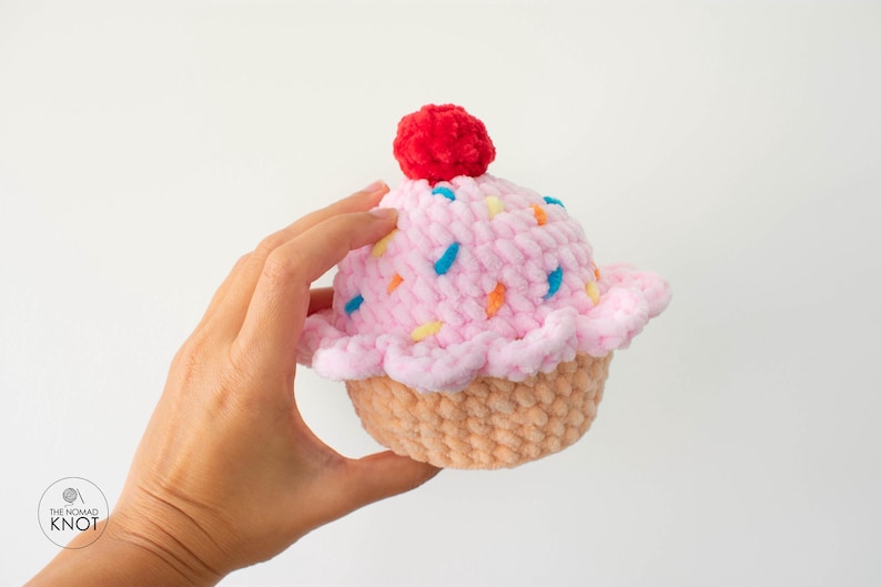 Op de afbeelding: Een handgemaakte gehaakte cupcake met roze glazuur, kleurrijke sprinkles en een rode kers. De cupcake heeft een lichtbruine, getextureerde basis en wordt in een hand gehouden tegen een witte achtergrond. Het Nomad Knot-logo staat linksonder.