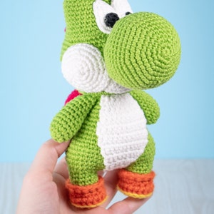 May include: Green crocheted Yoshi plush toy with white eyes, a white belly, and orange shoes. The toy is being held in a person's hand.