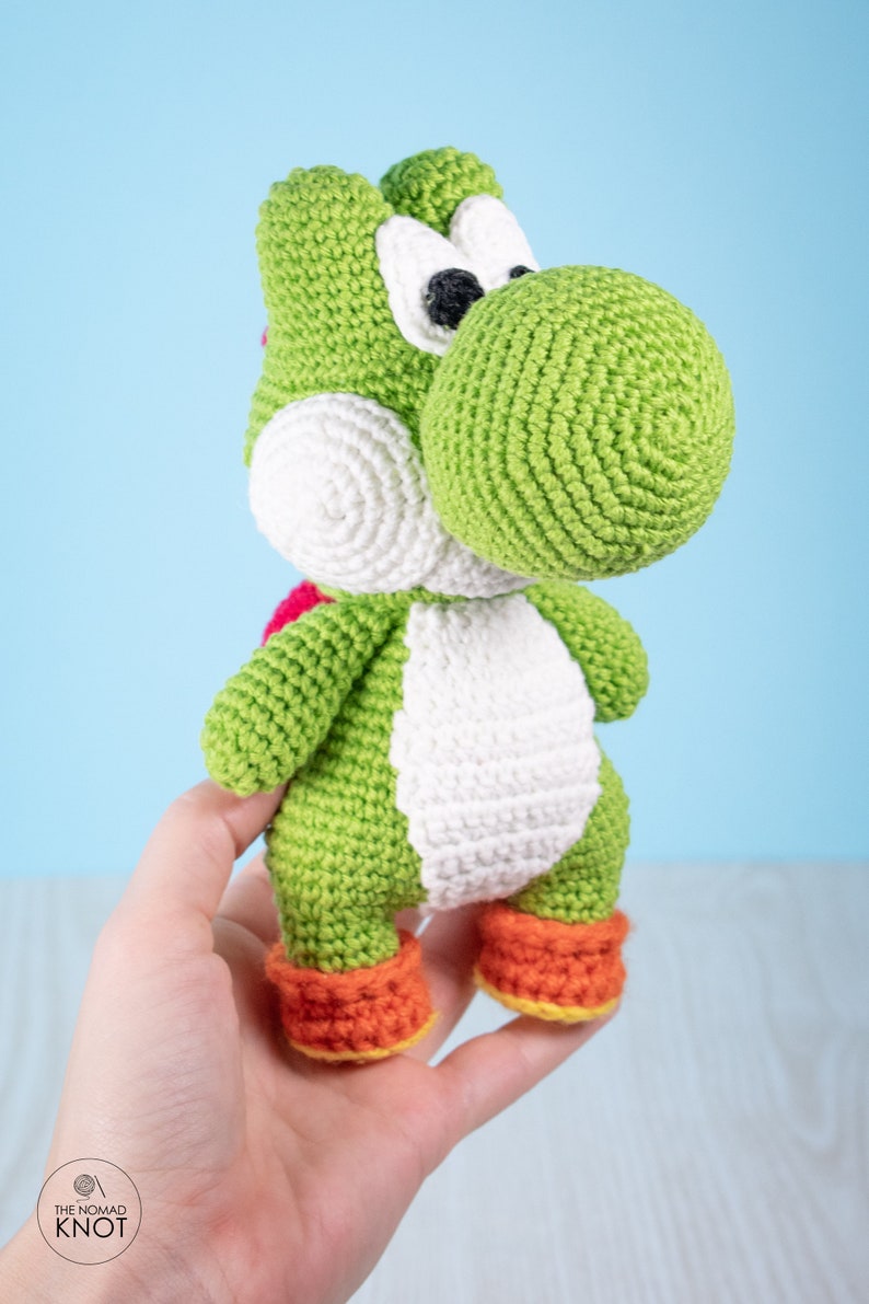 May include: Green crocheted Yoshi plush toy with white eyes, a white belly, and orange shoes. The toy is being held in a person's hand.