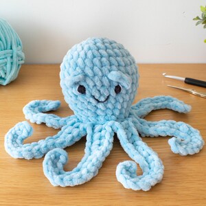 May include: A light blue crocheted octopus with a smiling face and eight curled tentacles. A ball of matching yarn sits to the left. A crochet hook and scissors are visible in the background. The Nomad Knot logo is in the lower left corner.
