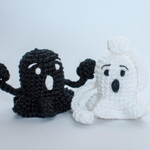 May include: Two crocheted ghost figures, one black and one white, with embroidered eyes and mouths. The black ghost is holding its arms out, while the white ghost is holding its hands up to its face.