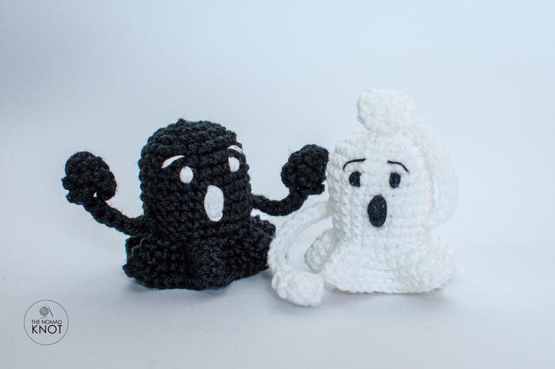 May include: Two crocheted ghost figures, one black and one white, with embroidered eyes and mouths. The black ghost is holding its arms out, while the white ghost is holding its hands up to its face.