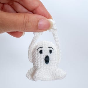 May include: A white crocheted ghost with black eyes and a mouth, hanging by its arms. The ghost is small and is being held by a hand.