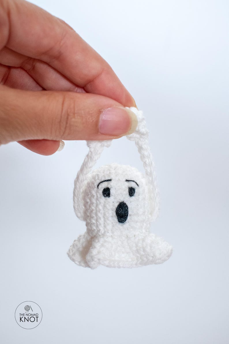 May include: A white crocheted ghost with black eyes and a mouth, hanging by its arms. The ghost is small and is being held by a hand.
