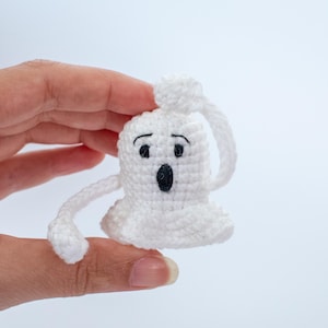 May include: A white crocheted ghost with black eyes and a mouth, held in a hand. The ghost has a long, thin arm extending to the left.