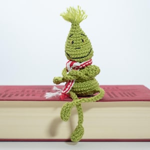 May include: A green crocheted creature with a red and white scarf sits on top of a closed book. The creature has a smiling face and long, thin legs.