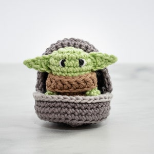 以下が含まれることがあります： グレーの編みぐるみポッドに座っている、緑と茶色の編みぐるみヨーダのキャラクター
