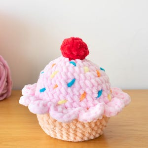 Op de afbeelding: Een handgemaakte, pluche cupcake met roze glazuur, versierd met kleurrijke sprinkles en een rode kers. De cupcake staat op een lichtbruine basis. Het logo van The Nomad Knot is zichtbaar.