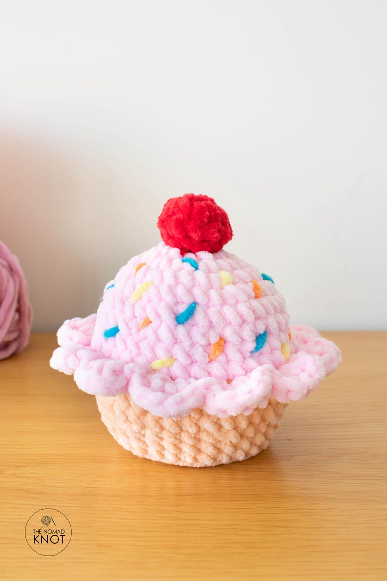 Op de afbeelding: Een handgemaakte, pluche cupcake met roze glazuur, versierd met kleurrijke sprinkles en een rode kers. De cupcake staat op een lichtbruine basis. Het logo van The Nomad Knot is zichtbaar.
