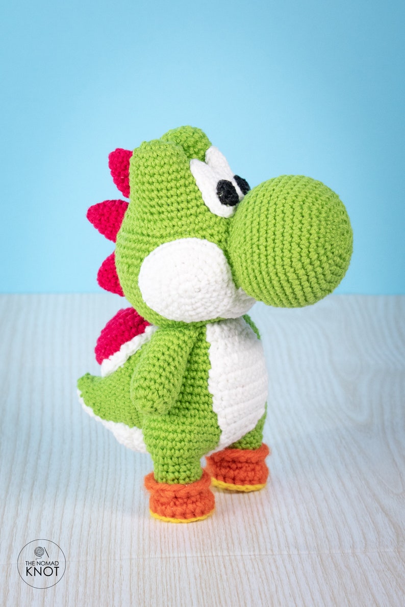 May include: Green crocheted Yoshi plush toy with white eyes, a red spiky back, and orange shoes. The toy is standing on a light wood surface.