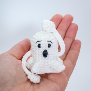 May include: A white crocheted ghost with black eyes and a mouth, held in a hand. The ghost has a long tail that is also crocheted.