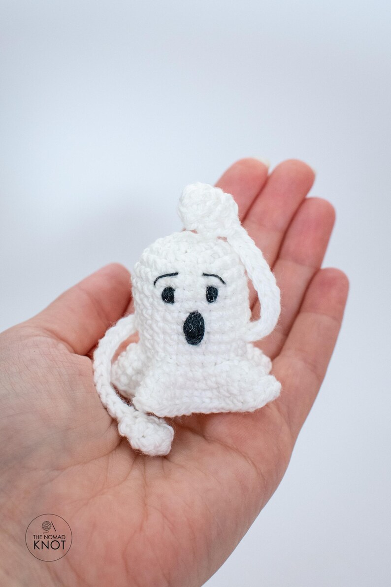 May include: A white crocheted ghost with black eyes and a mouth, held in a hand. The ghost has a long tail that is also crocheted.