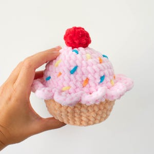 Op de afbeelding: Een handgemaakte pluche cupcake met roze glazuur en kleurrijke hagelslag. Een rode pompon bedekt de cupcake. De basis is lichtbruin. Het logo The Nomad Knot is linksonder zichtbaar.