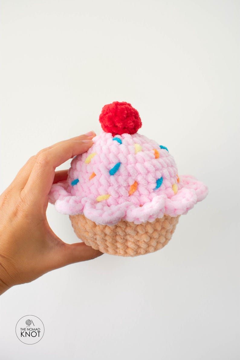 Op de afbeelding: Een handgemaakte pluche cupcake met roze glazuur en kleurrijke hagelslag. Een rode pompon bedekt de cupcake. De basis is lichtbruin. Het logo The Nomad Knot is linksonder zichtbaar.
