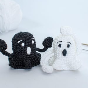 May include: Two crocheted ghost figures, one black and one white, with white eyes and mouths. The ghosts are standing on a white surface.