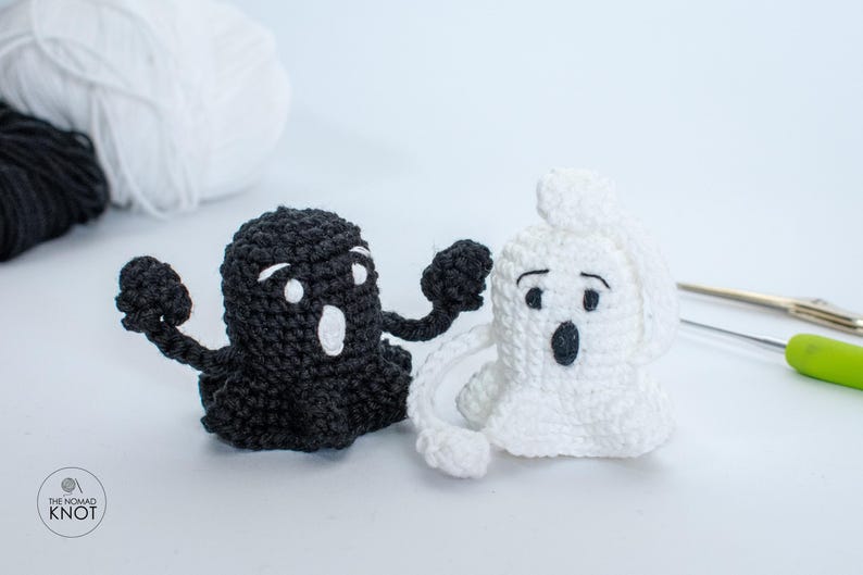 May include: Two crocheted ghost figures, one black and one white, with white eyes and mouths. The ghosts are standing on a white surface.