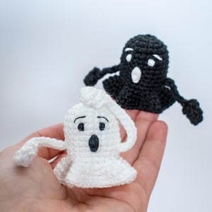 May include: Two crocheted ghost finger puppets, one white and one black, with black eyes and mouths. The puppets are held in a hand against a white background.