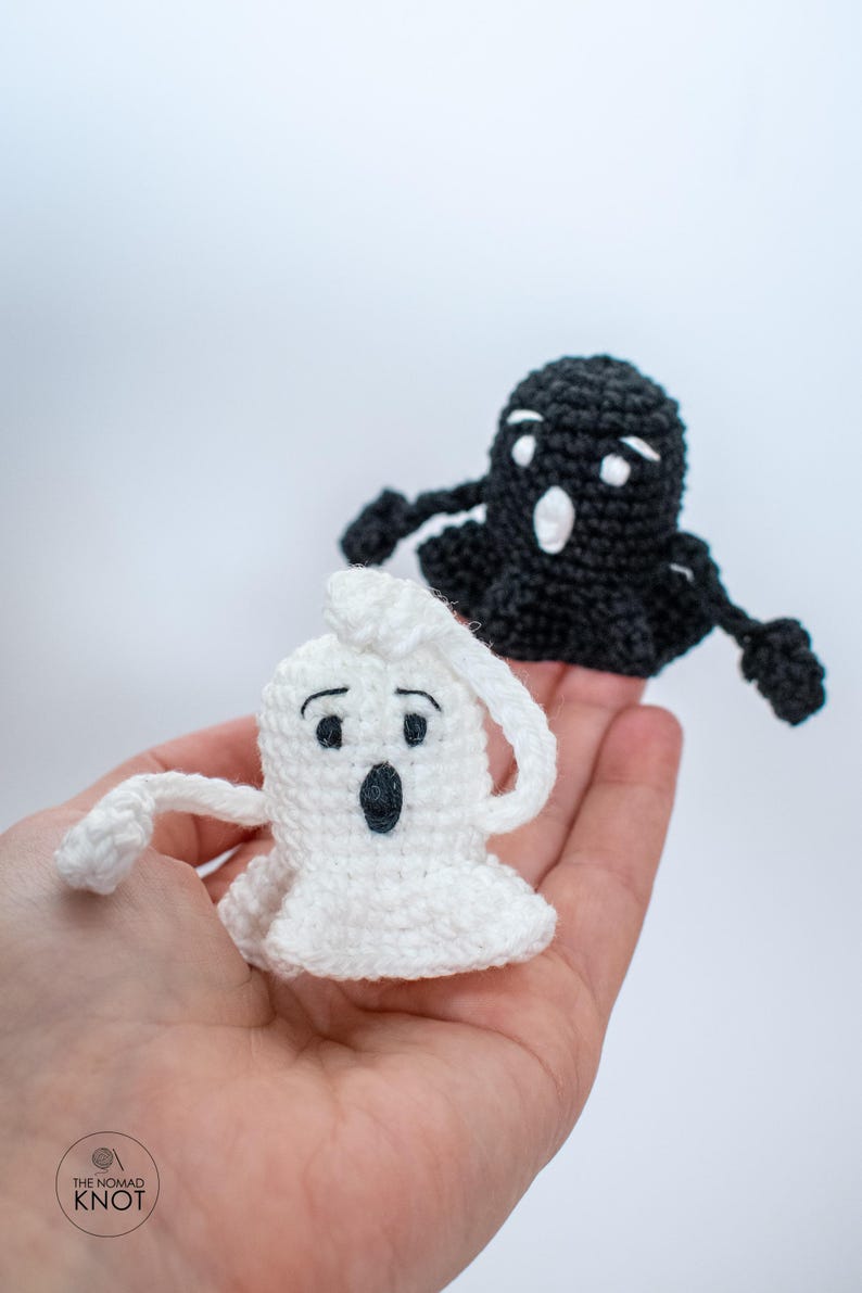 May include: Two crocheted ghost finger puppets, one white and one black, with black eyes and mouths. The puppets are held in a hand against a white background.