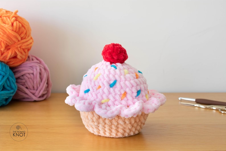 Op de afbeelding: Een handgemaakte gehaakte cupcake met roze glazuur, kleurrijke sprinkles en een rode kers. De cupcake staat op een lichtbruine basis. Bollen oranje, roze en blauw garen op de achtergrond. Een schaar en een haaknaald rechts.