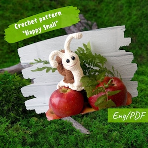 Puede incluir: Patrón de crochet para un amigurumi "Happy Snail". El caracol es blanco con detalles marrones y está sentado sobre dos manzanas rojas. El patrón está disponible en inglés y en formato PDF.