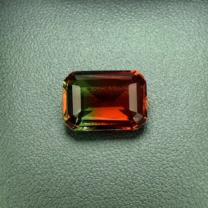 May include: A green, red, and orange gemstone in a rectangular shape is displayed in a black velvet box.