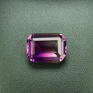 May include: A purple gemstone with a rectangular emerald cut, displayed in a black leather ring box.