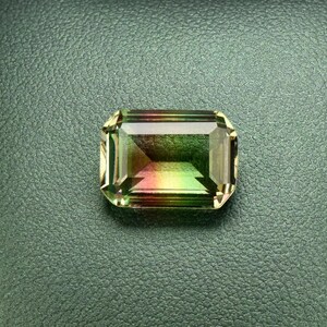 May include: A green, yellow, and pink gemstone with a rectangular shape is displayed in a black leather box.