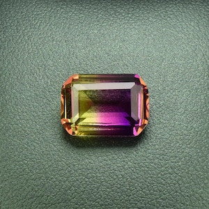 May include: A vibrant, multi-colored gemstone with a rectangular shape, set in a black leather display box. The gemstone has a gradient of yellow, orange, and purple hues.