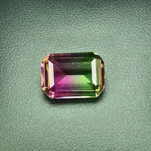 May include: A green, pink, and yellow gemstone in a rectangular shape is displayed in a black leather box.