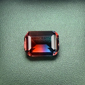 May include: A red, blue, and green gemstone in an emerald cut setting, displayed in a black velvet jewelry box.