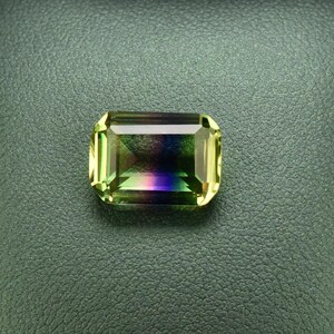 May include: A green, purple, and yellow gemstone in a square emerald cut, set in a black leather ring box.