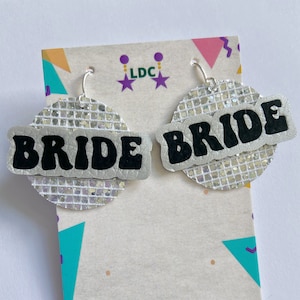 May include: A pair of silver glitter earrings with black text that reads "BRIDE". The earrings are on a white card with a colourful geometric pattern and the text "ldcstore.uk".