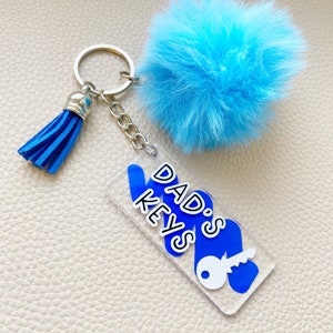 May include: Blue and white keyring with a blue pom-pom charm and a tassel. The keyring has a clear acrylic tag with the text "DAD'S KEYS" and a key icon.
