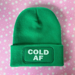 May include: A green beanie with a white patch that says "COLD AF" on it.