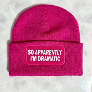May include: A vibrant pink beanie hat with a turned-up brim. The hat has a rectangular patch with the white text "SO APPARENTLY I'M DRAMATIC".